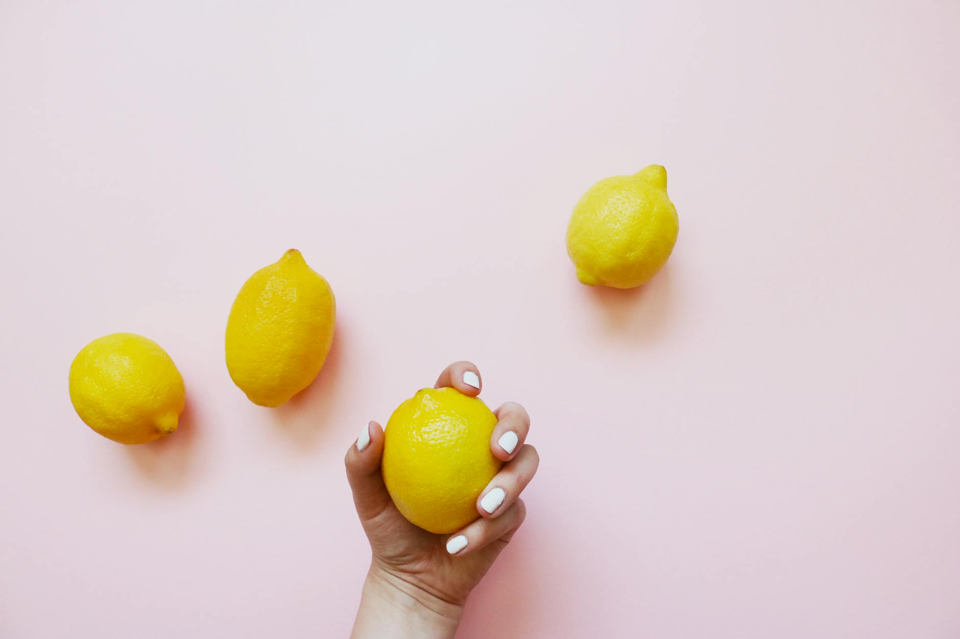 A hand holding a lemon on a soft pink background surrounded by fresh lemons.