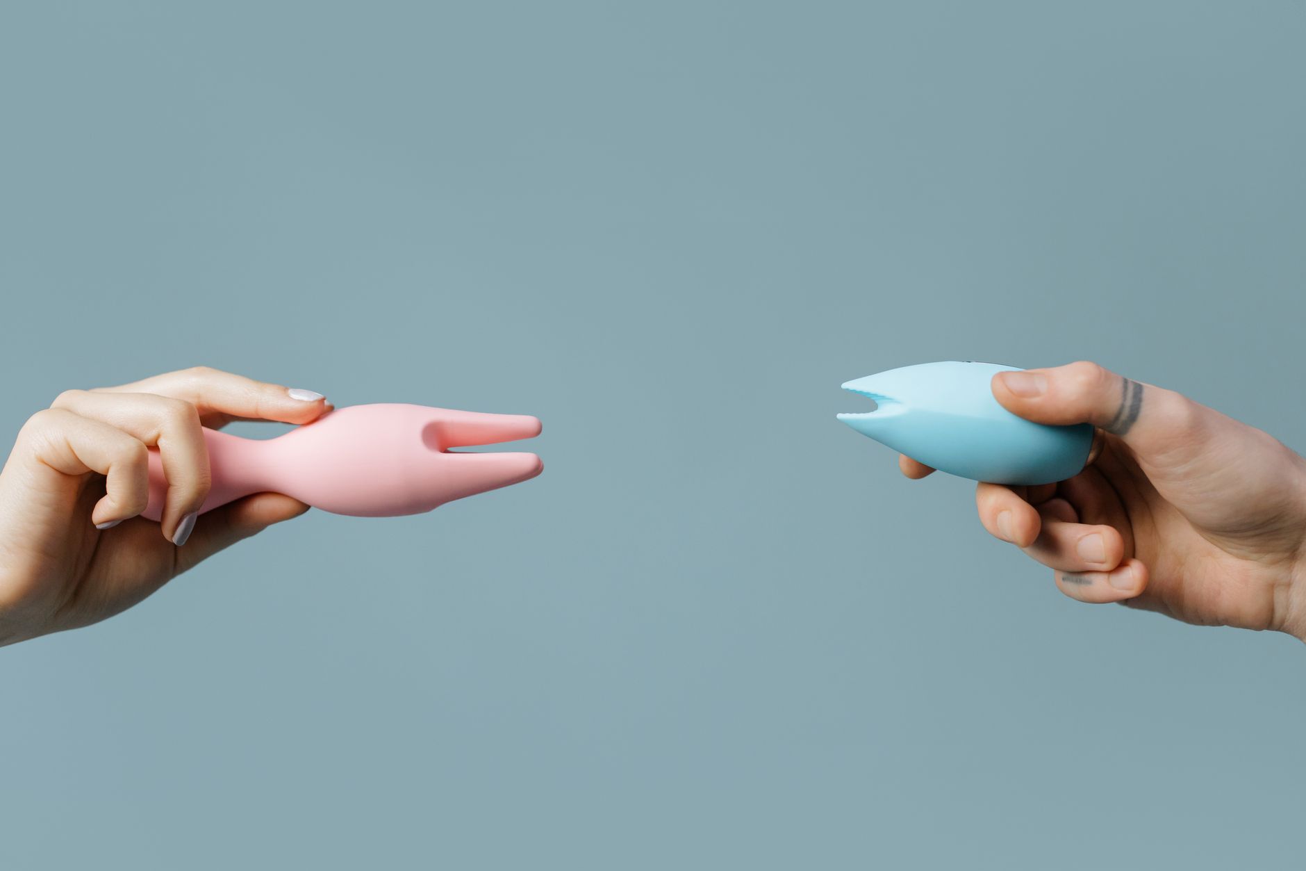 Two hands holding pink and blue silicone clitoral vibrators against a pastel background