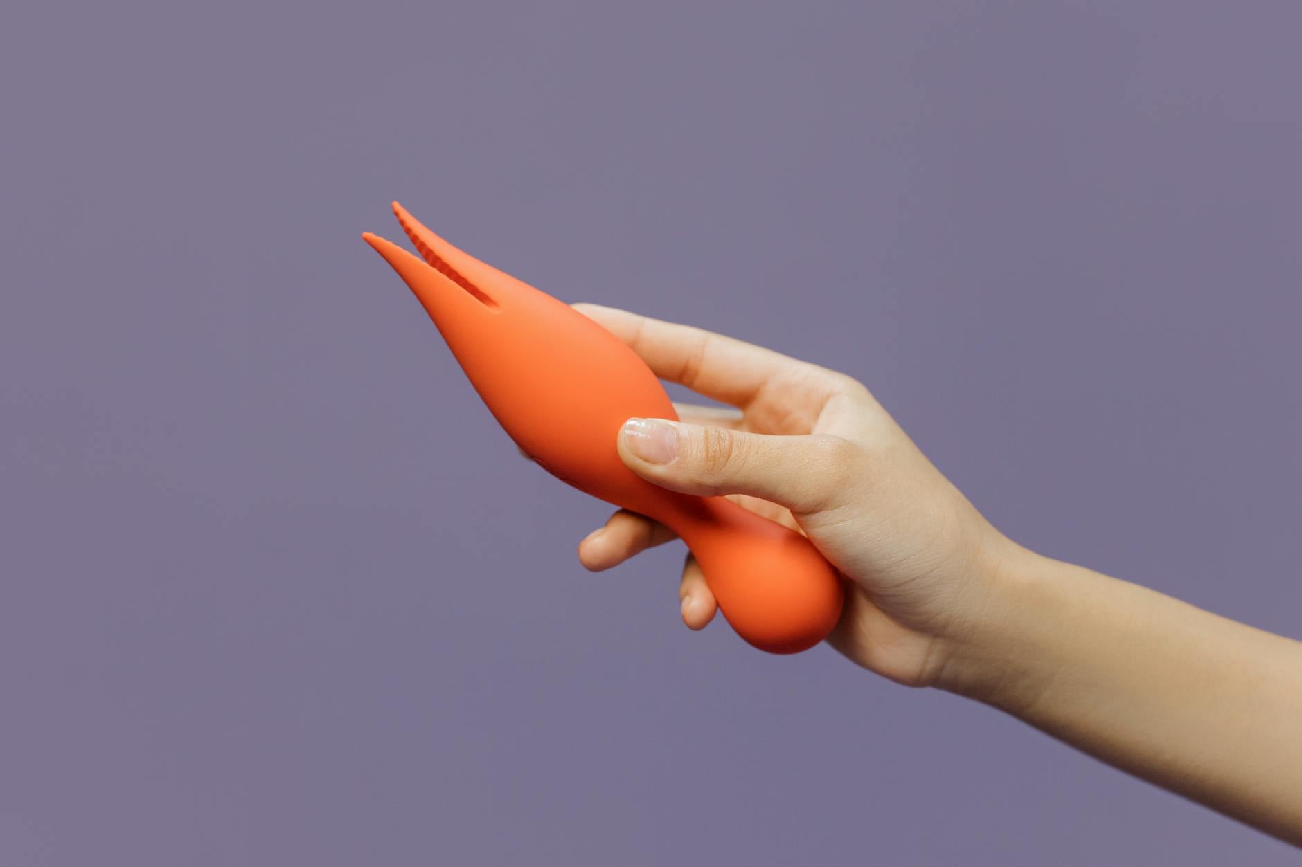 A hand holding an orange vibrator against a minimalistic purple backdrop, symbolizing modern solo and partnered pleasure