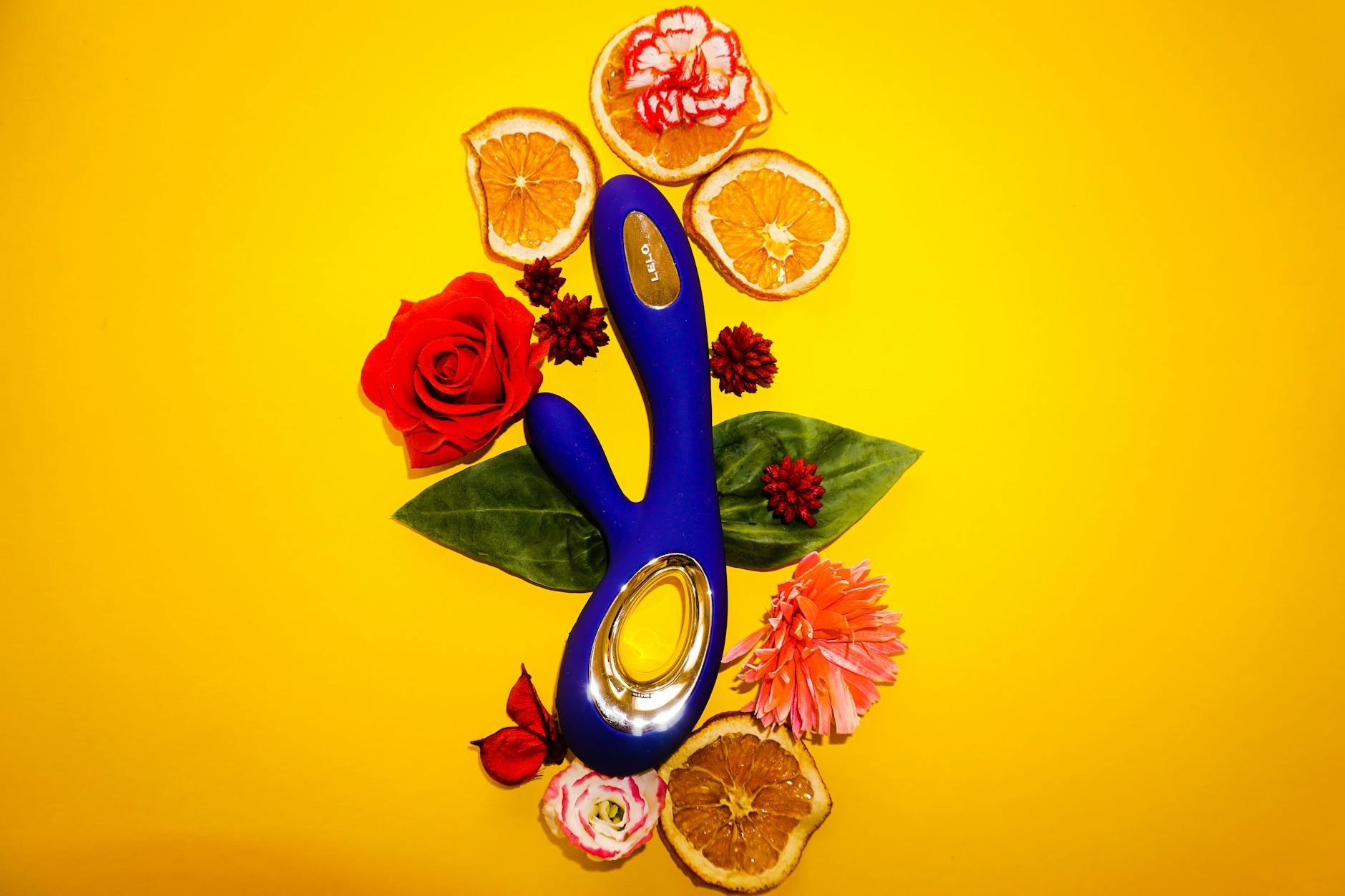 A blue clitoral vibrator surrounded by fresh citrus fruits and flowers on a bright yellow background